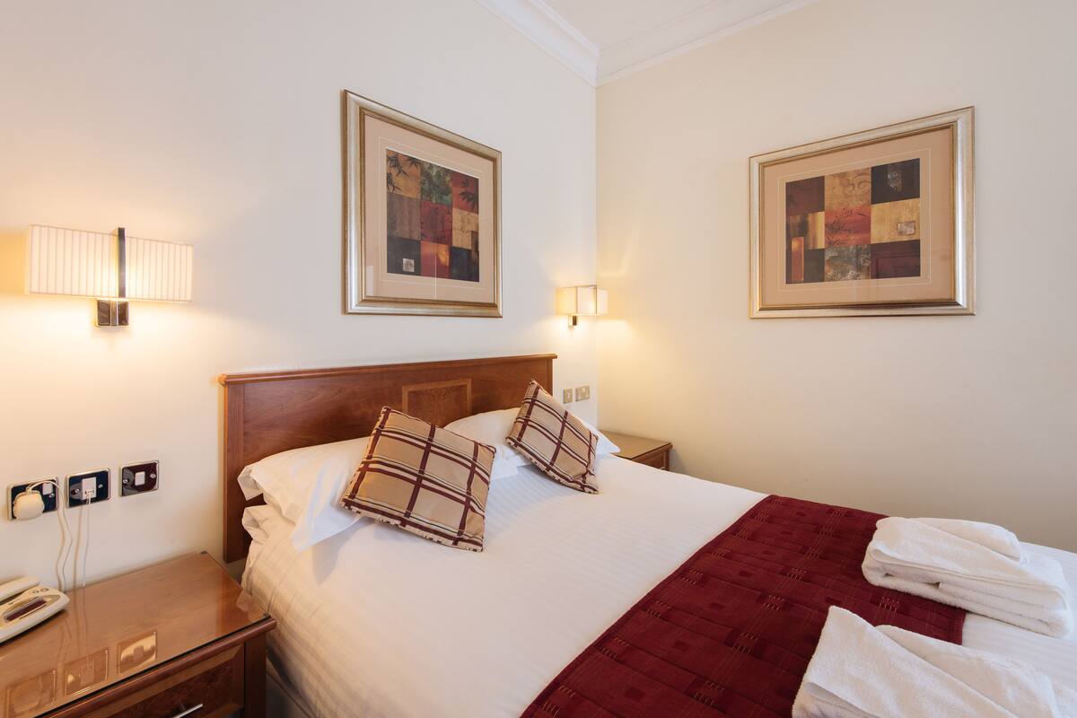 A hotel room with a neatly made bed, two framed artworks on the walls, a bedside lamp, a telephone, and a red throw blanket.