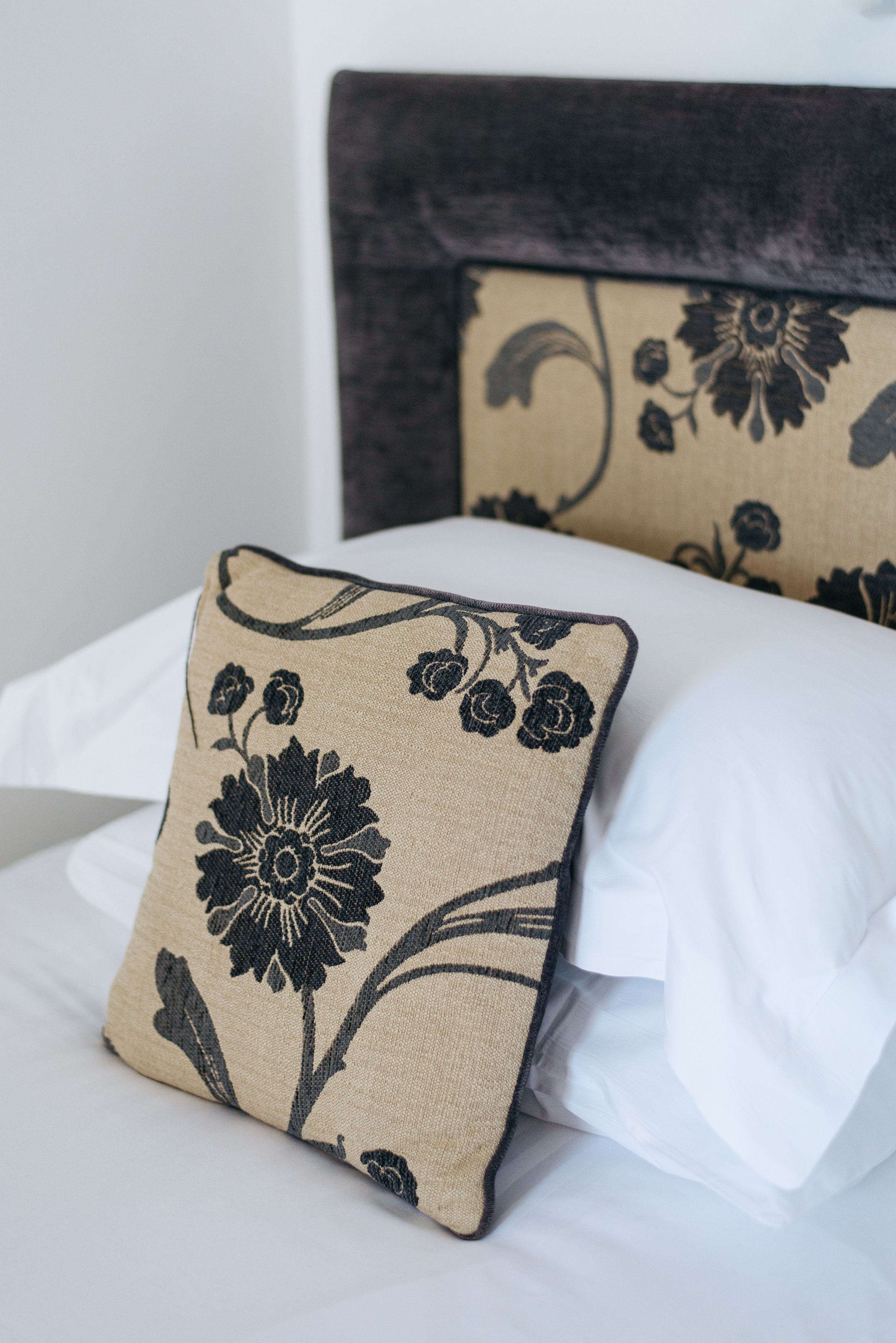 Una cama con sábanas blancas y un almohadón color beige con estampado floral negro.