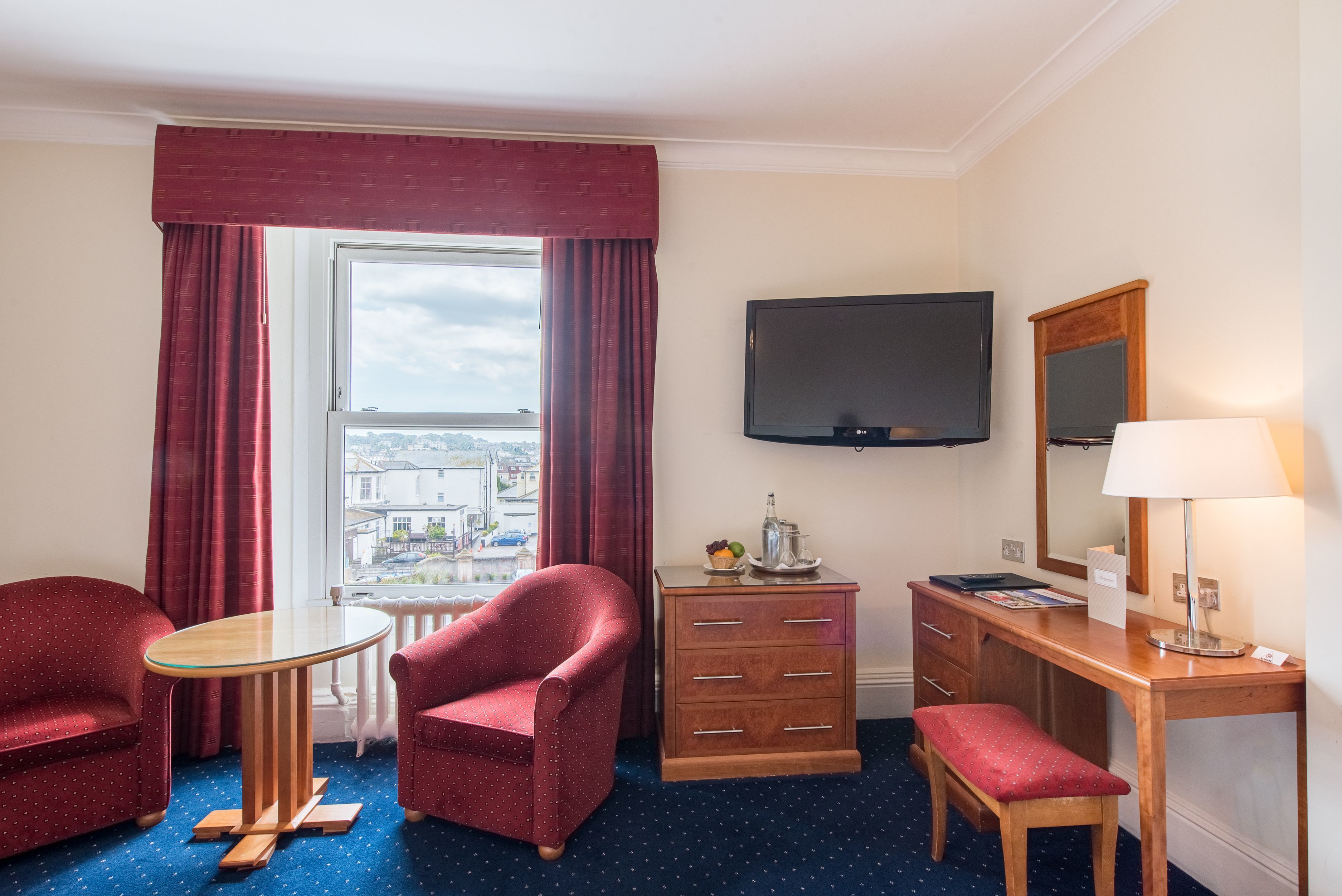 A hotel room with a large window, a flat-screen TV, a wooden desk, a chair, a small table, and a bench.