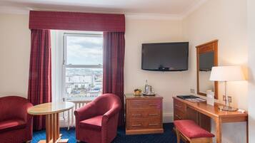 A hotel room with a large window, a flat-screen TV, a wooden desk, a chair, a small table, and a bench.