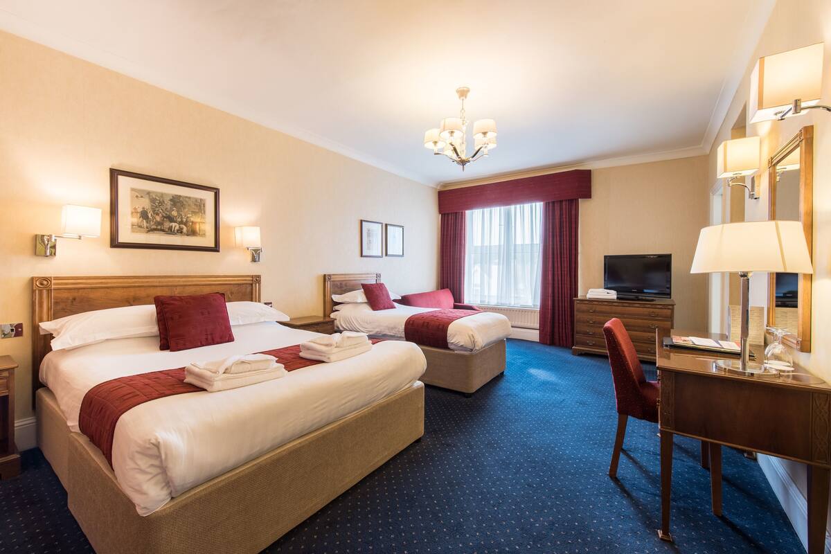 A hotel room with two beds, a desk, a chair, a television, and a chandelier.
