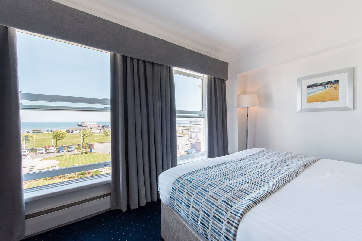 A hotel room with a large window, a bed with a patterned bedspread, a lamp, and a framed picture on the wall.