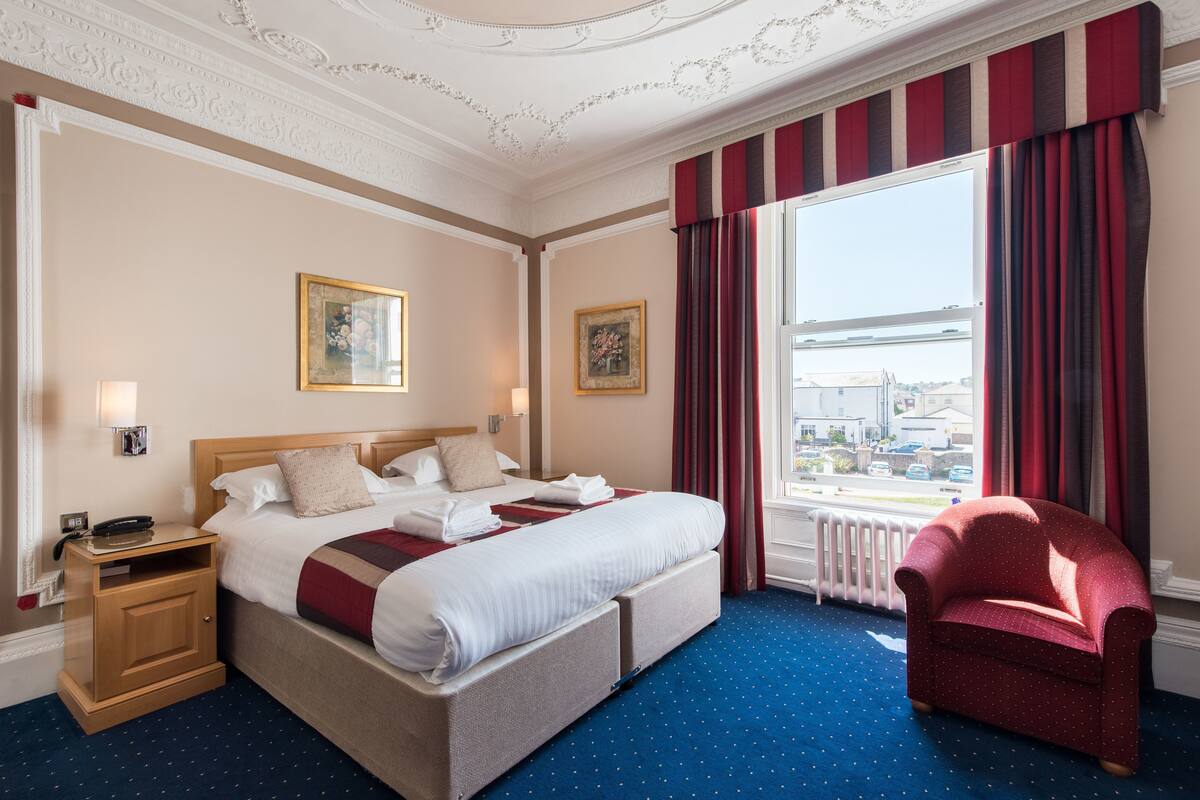A hotel room with a large bed, a wooden nightstand, a red armchair, and a window with curtains.
