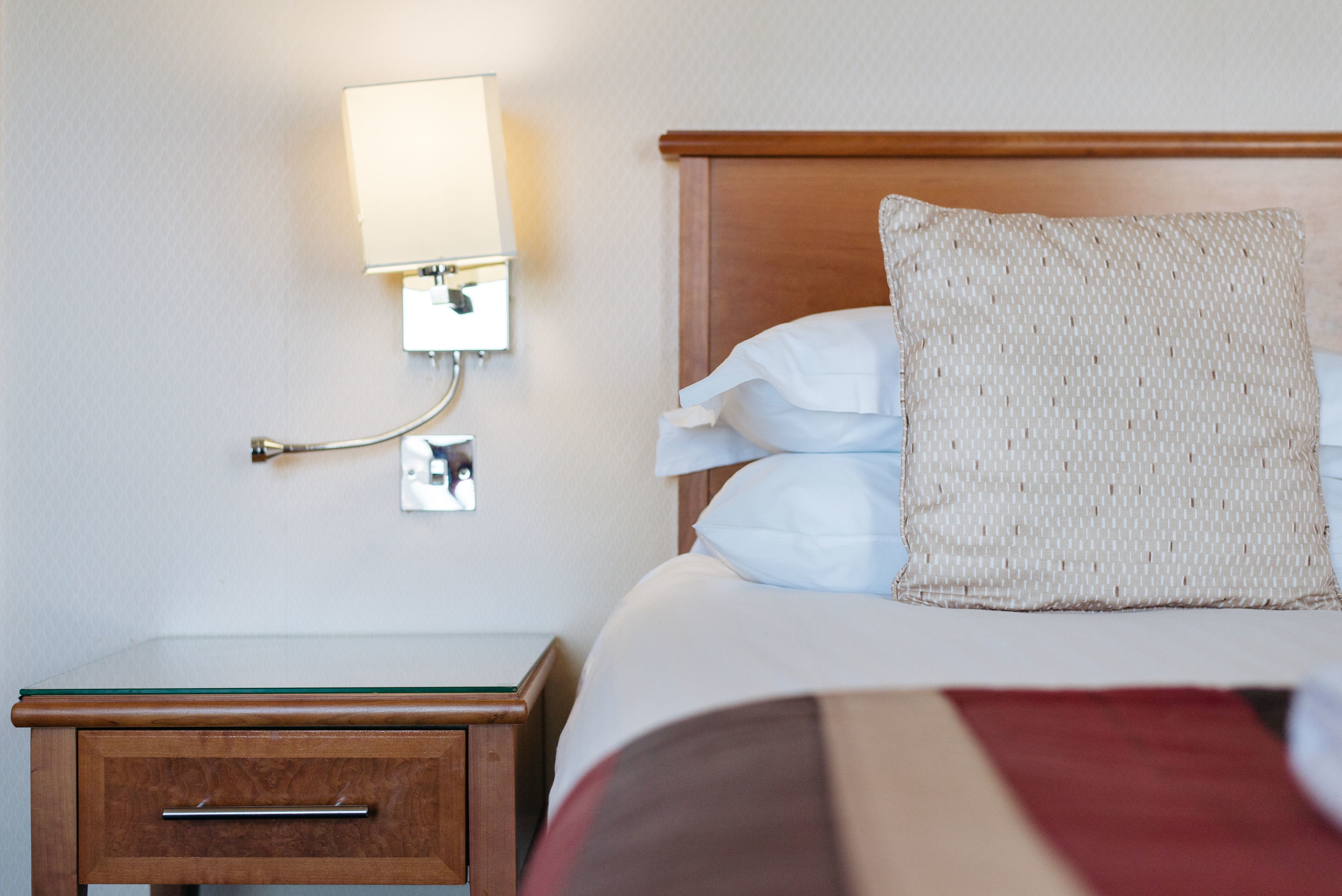 A neatly made bed with white linens and a woven pillow, a bedside table with a lamp, and a wall-mounted light fixture.