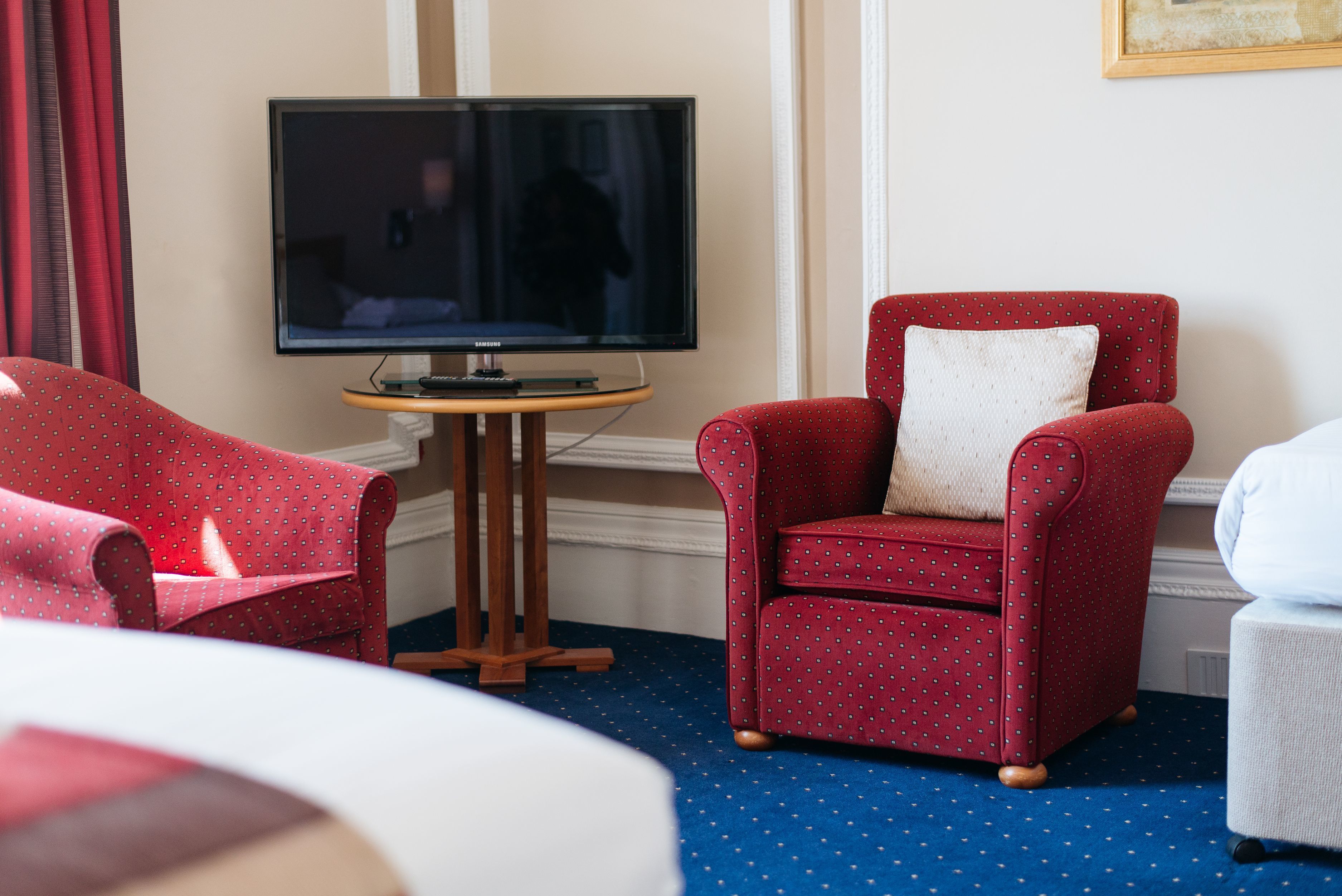 A hotel room with a large flat-screen TV on a wooden stand, two red upholstered chairs with a polka dot pattern, a white chair, and a blue carpet with white dots.