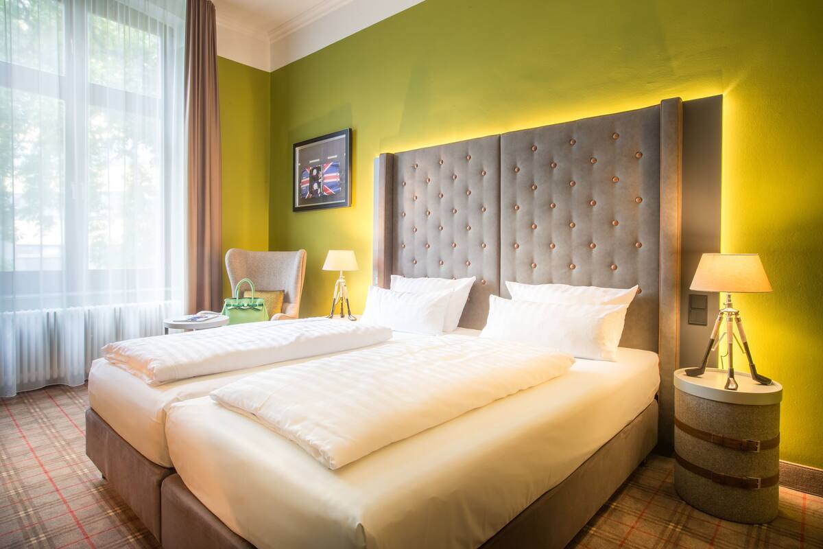 A double bed with a tufted headboard, two bedside tables with lamps, a green wall, and a window with sheer curtains.