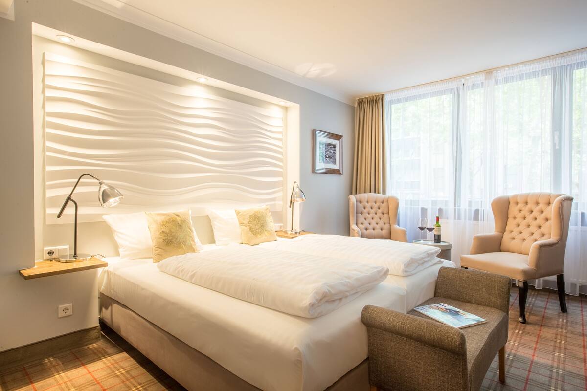 A hotel room with a large bed, two armchairs, a bedside table with a lamp, and a window with curtains.