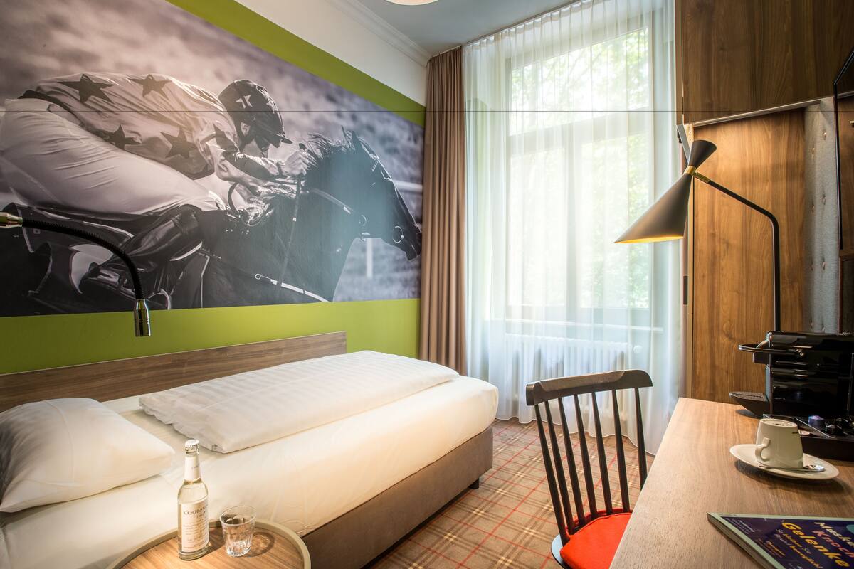 A hotel room with a bed, a desk with a chair, a lamp, a coffee cup, and a bottle on a side table.