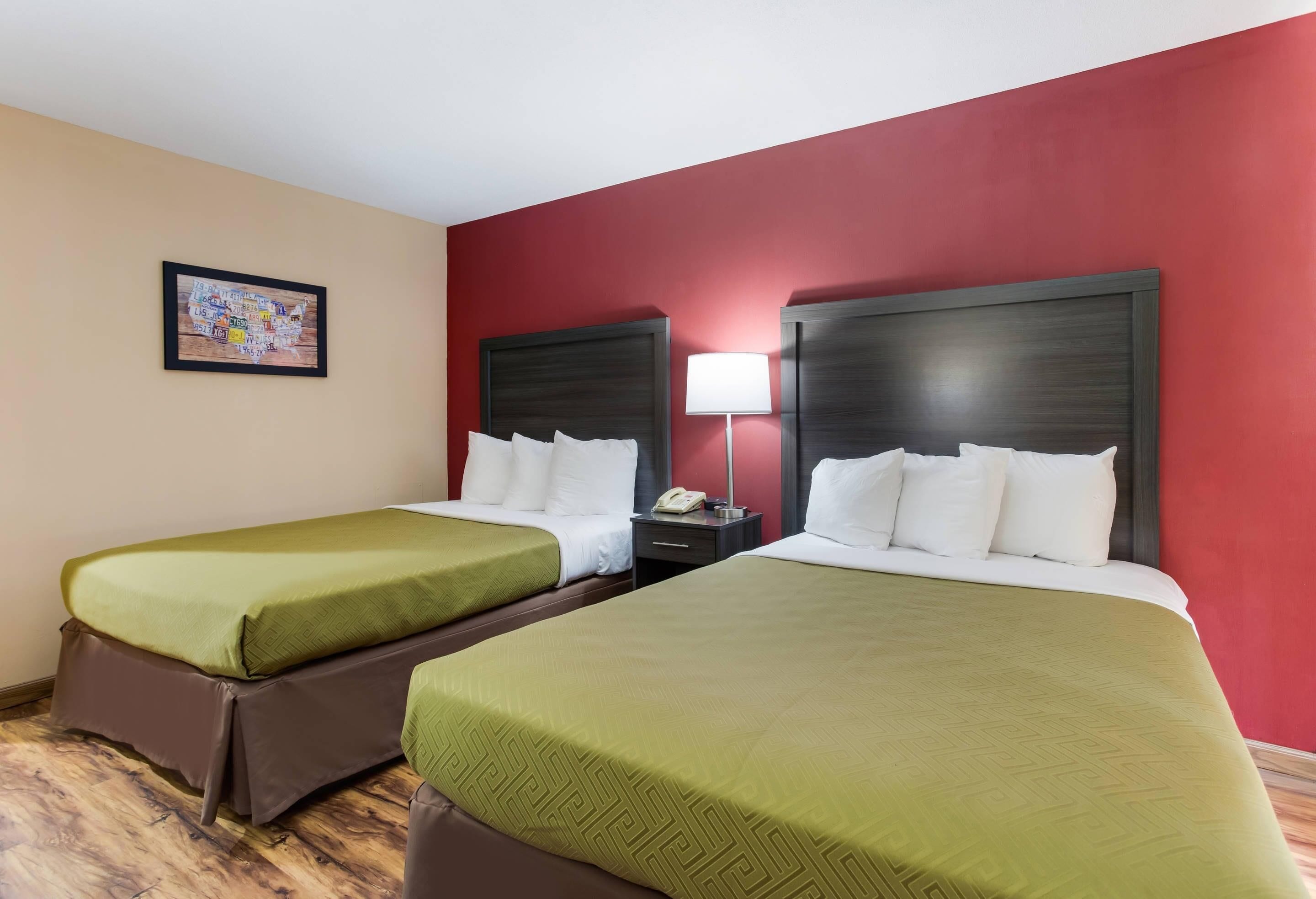A hotel room with two beds, each with green bedding and white pillows, a bedside table with a lamp, and a framed picture on the wall.