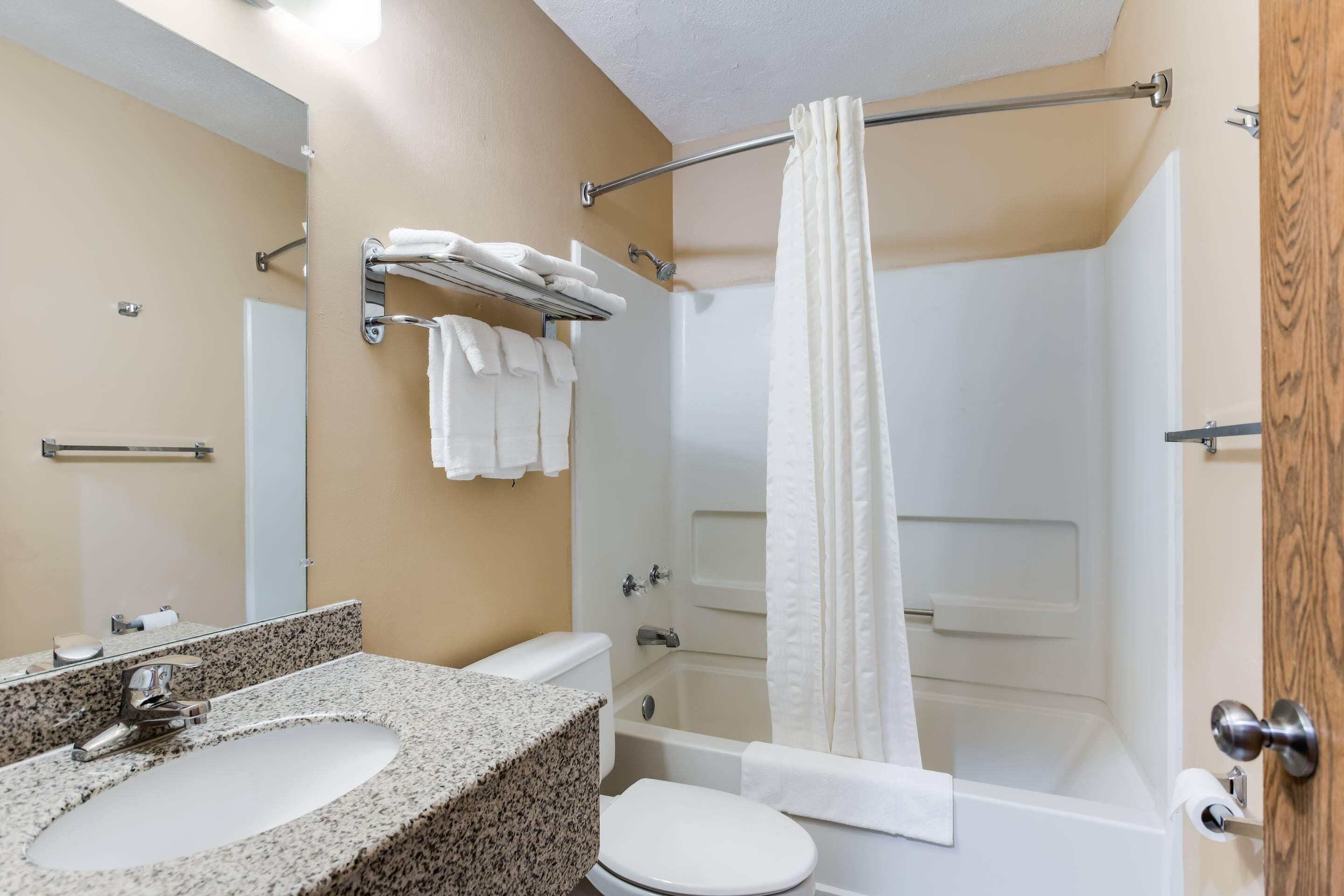 Un baño con lavamanos de encimera de granito, inodoro blanco y ducha a pie con cortina blanca.