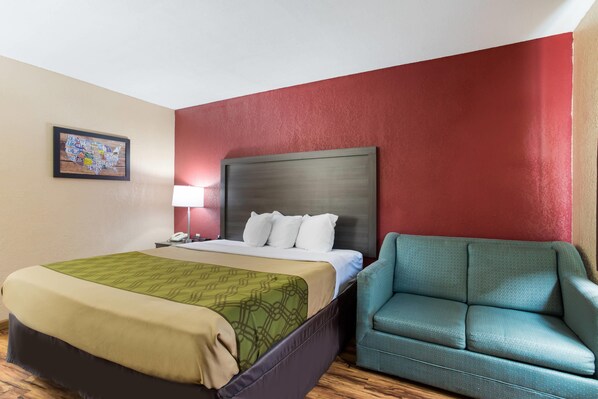 A hotel room with a bed, a green patterned bedspread, a teal armchair, a red accent wall, and a framed map on the wall.