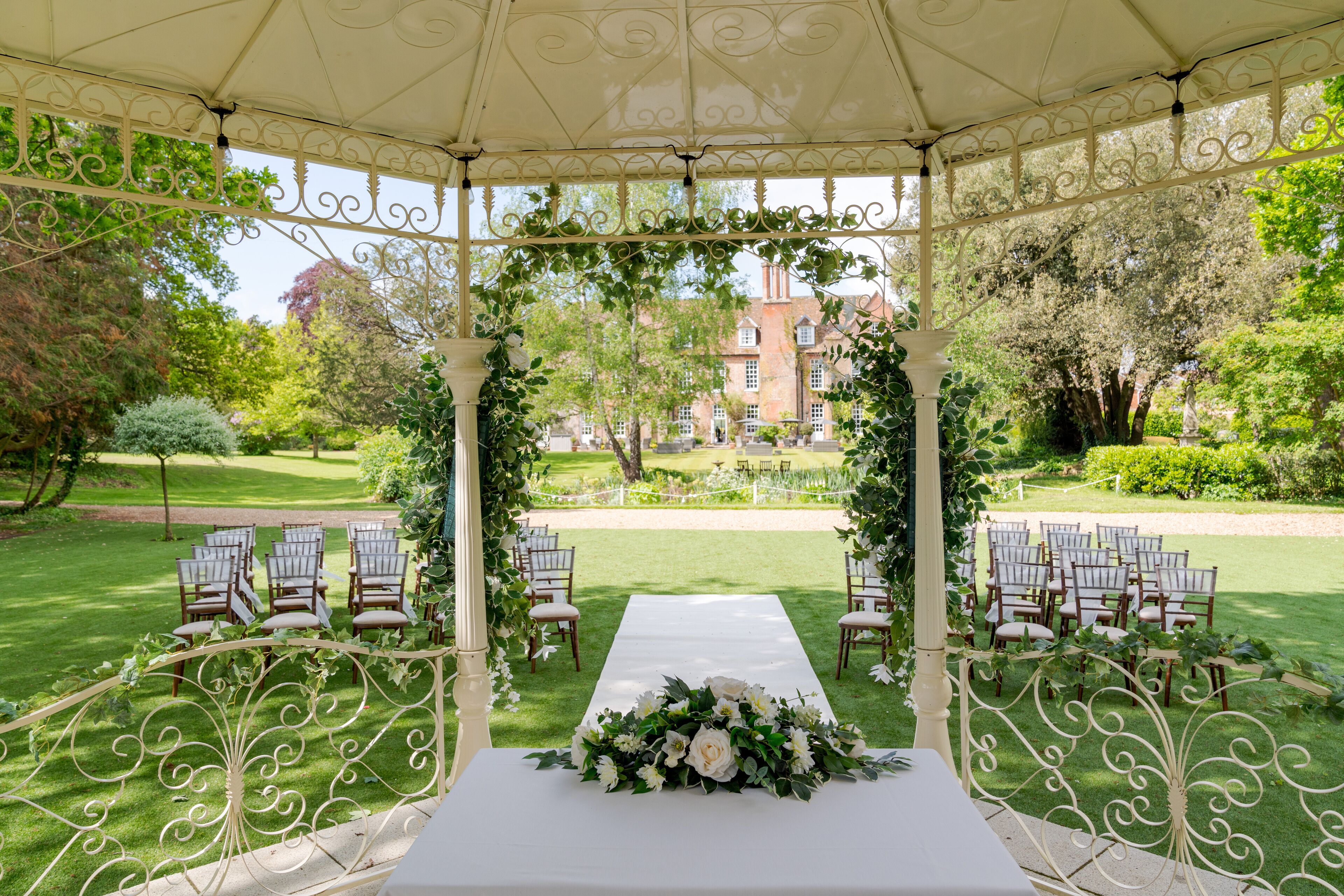 A gazebo with a white table and chairs, surrounded by greenery and a lawn.