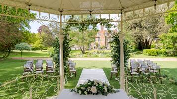A gazebo with a white table and chairs, surrounded by greenery and a lawn.