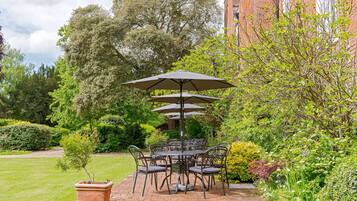 A patio with a table and chairs, surrounded by lush greenery and a brick building in the background.