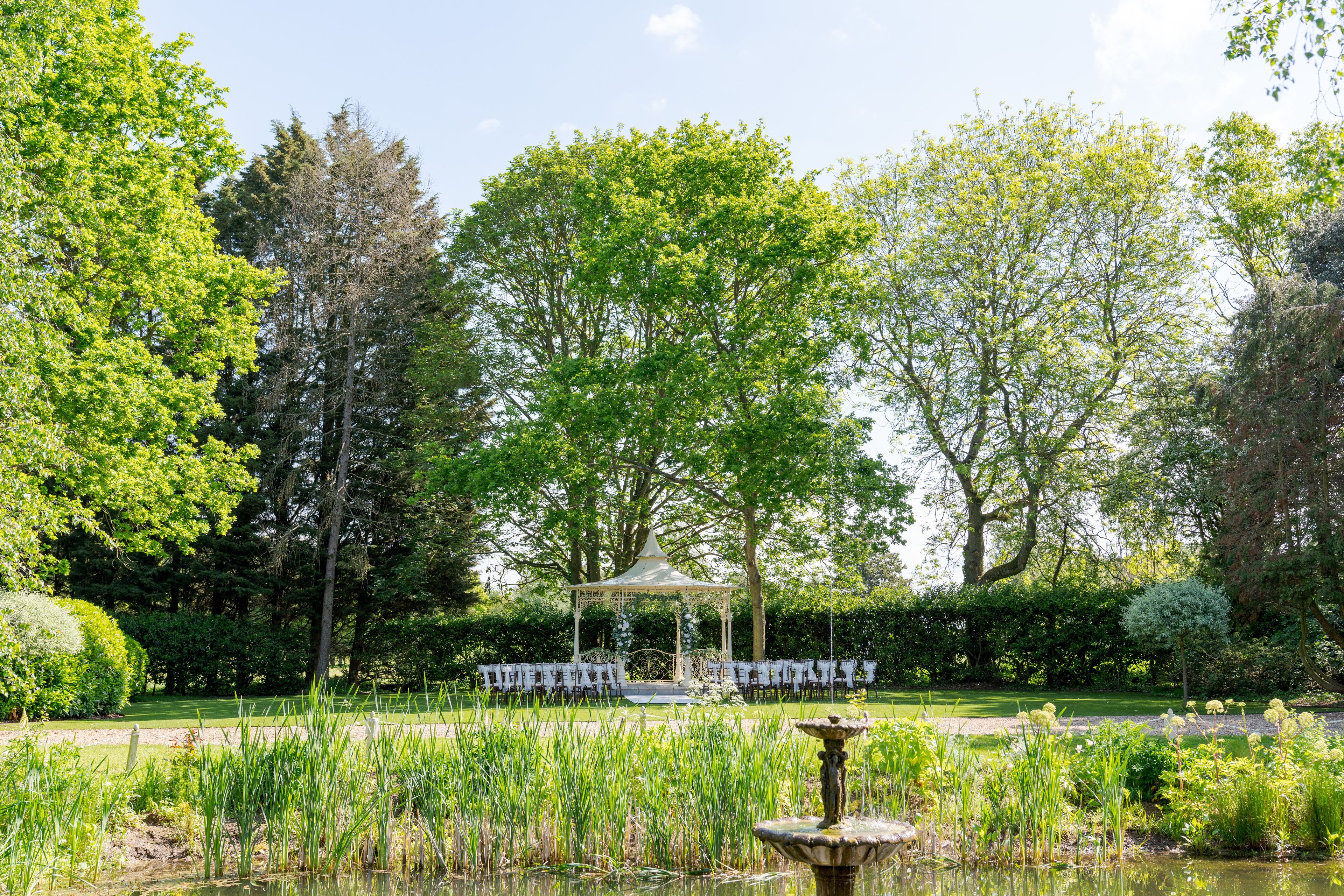 A gazebo with a white canopy, surrounded by a garden with a pond and various plants.