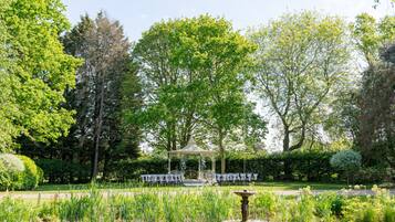 A gazebo with a white canopy, surrounded by a garden with a pond and various plants.