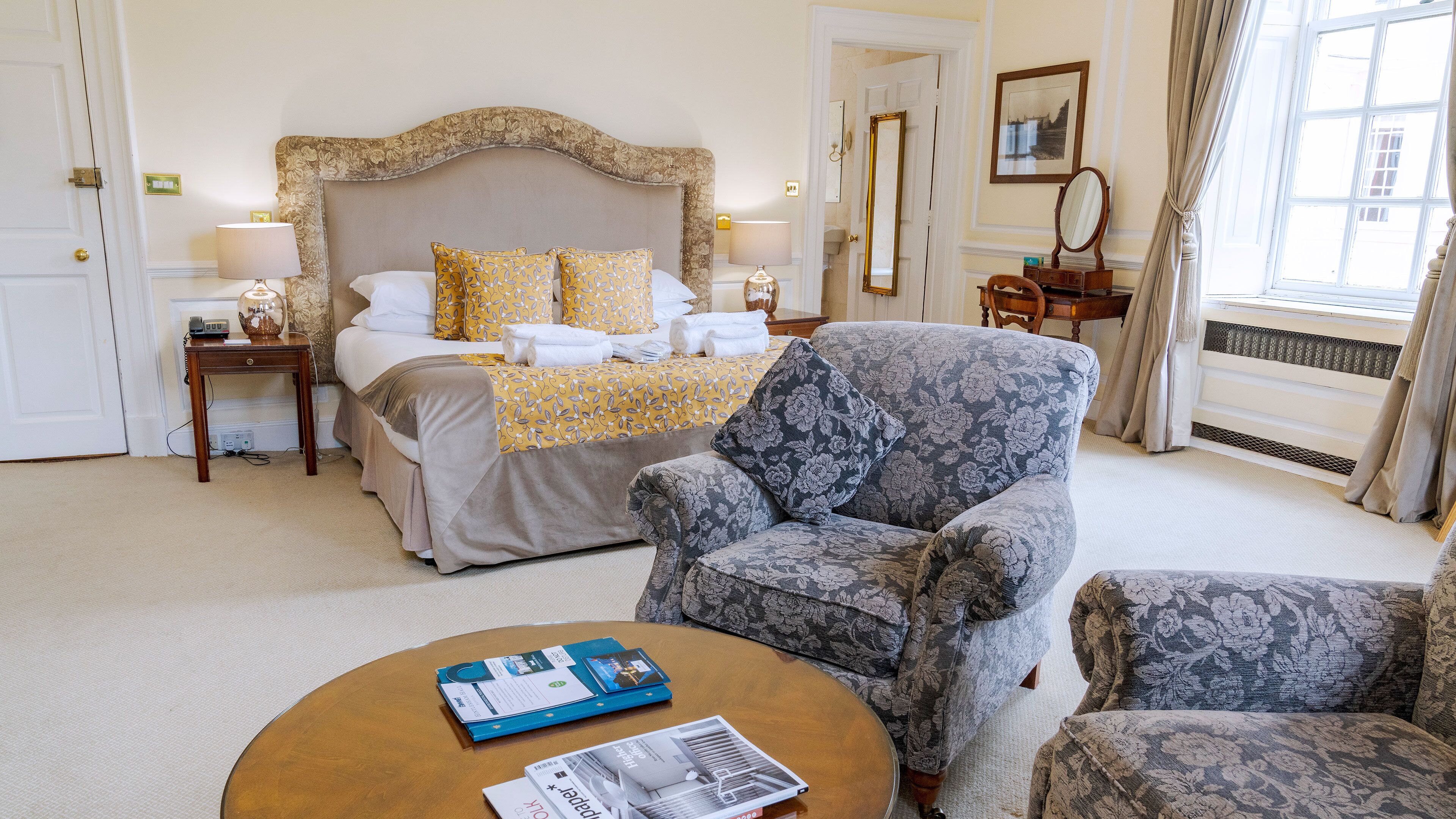 Un cuarto de hotel con una cama grande, un sillón floral, una mesita de centro redonda con revistas y vistas al exterior por una ventana.