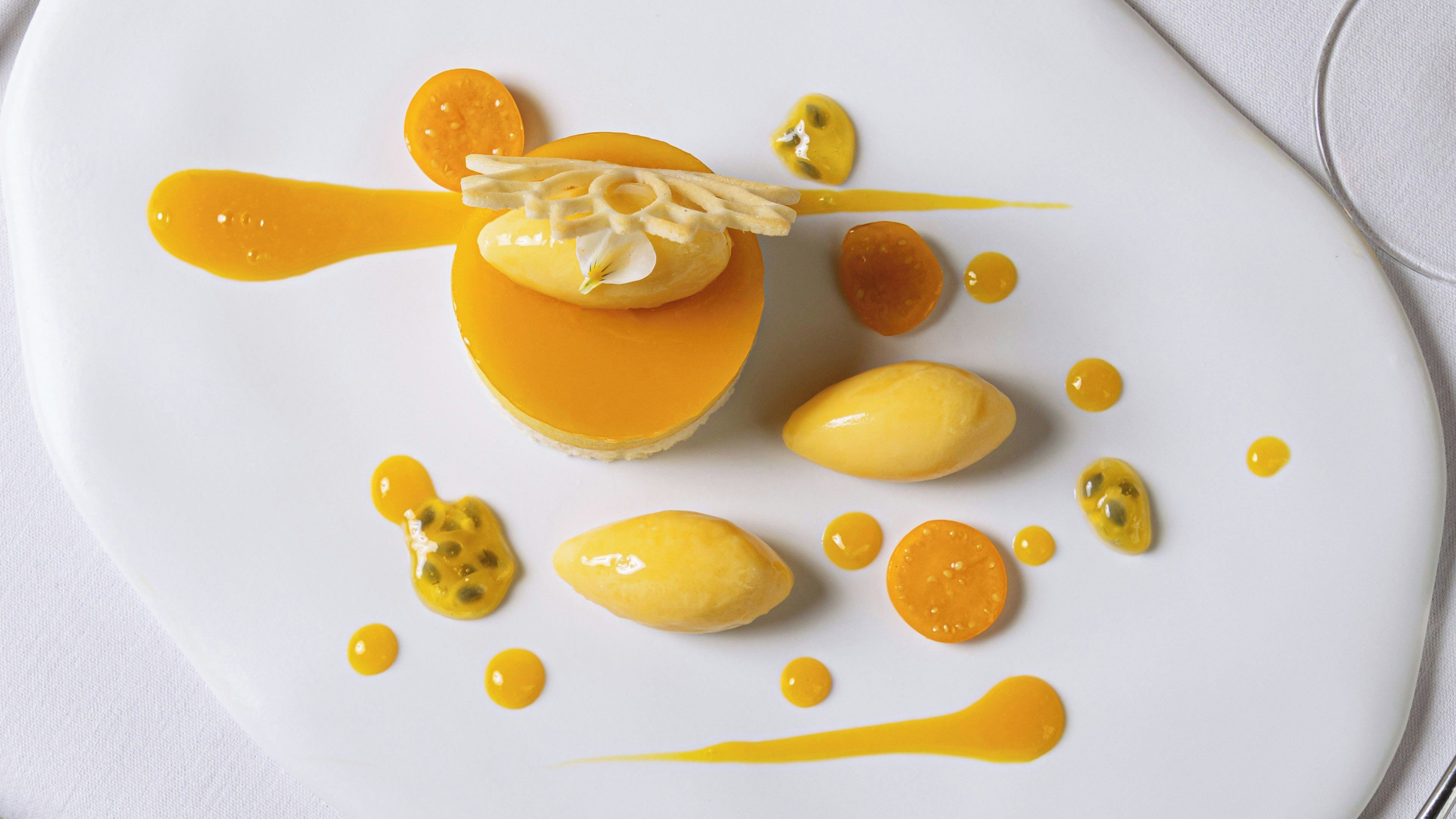 A dessert with a smooth, glossy topping and a dollop of cream on top, surrounded by small dollops of a yellow sauce and slices of a yellow fruit.
