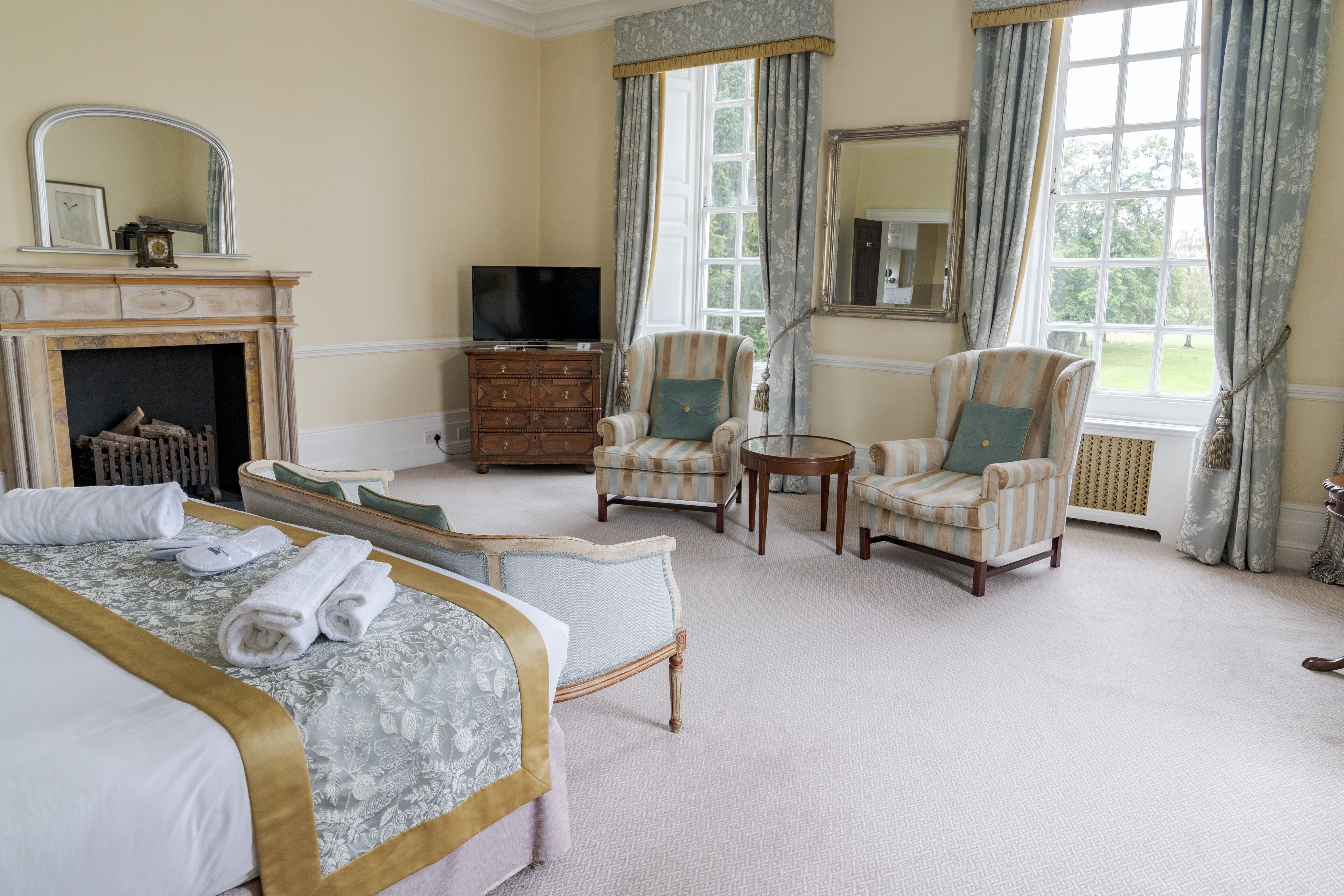 A hotel room with a bed, two armchairs, a small table, a TV, a fireplace, and large windows with curtains.