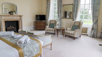 A hotel room with a bed, two armchairs, a small table, a TV, a fireplace, and large windows with curtains.