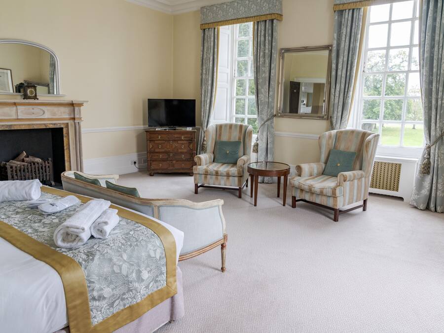 A hotel room with a bed, two armchairs, a small table, a TV, a fireplace, and large windows with curtains.