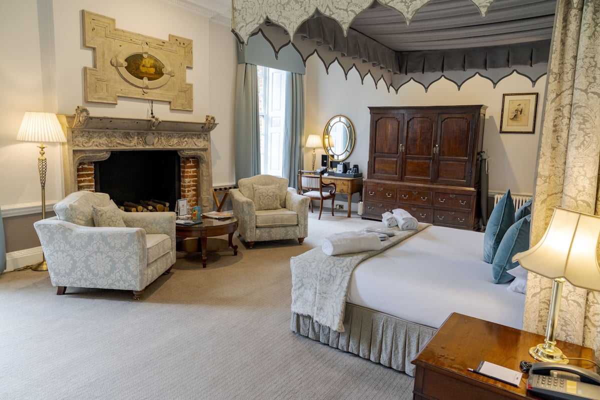 A hotel room with a fireplace, a bed, two armchairs, a desk with a mirror, and a wardrobe.