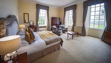 A spacious bedroom with a large bed, two armchairs, a wooden dresser, and a view of the outdoors through large windows.