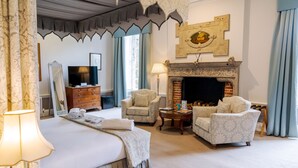 A hotel room with a bed, two armchairs, a small table, a fireplace, a television, and a mirror. - Hintlesham Hall Hotel (Ipswich)