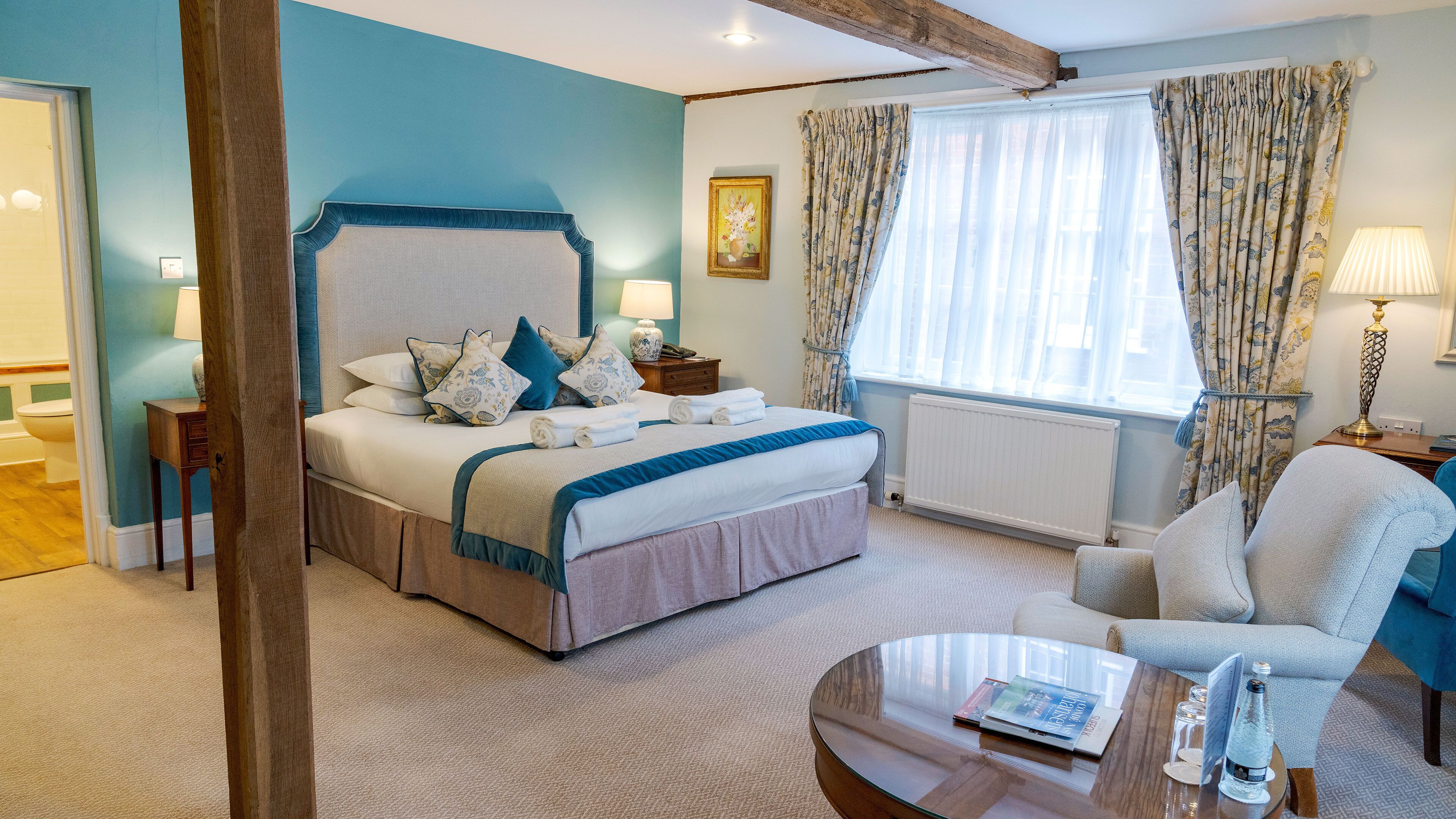 A bedroom with a large bed, a bedside table with a lamp, a window with curtains, and a chair.