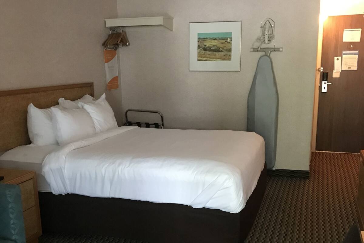 A hotel room with a large bed, a bedside table, a lamp, a picture on the wall, and a door with a light on.