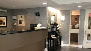 Uma área de recepção com balcão, máquina de café e lixeira. Há quadros com certificados na parede e um sinal indicando acessibilidade para cadeiras de rodas.