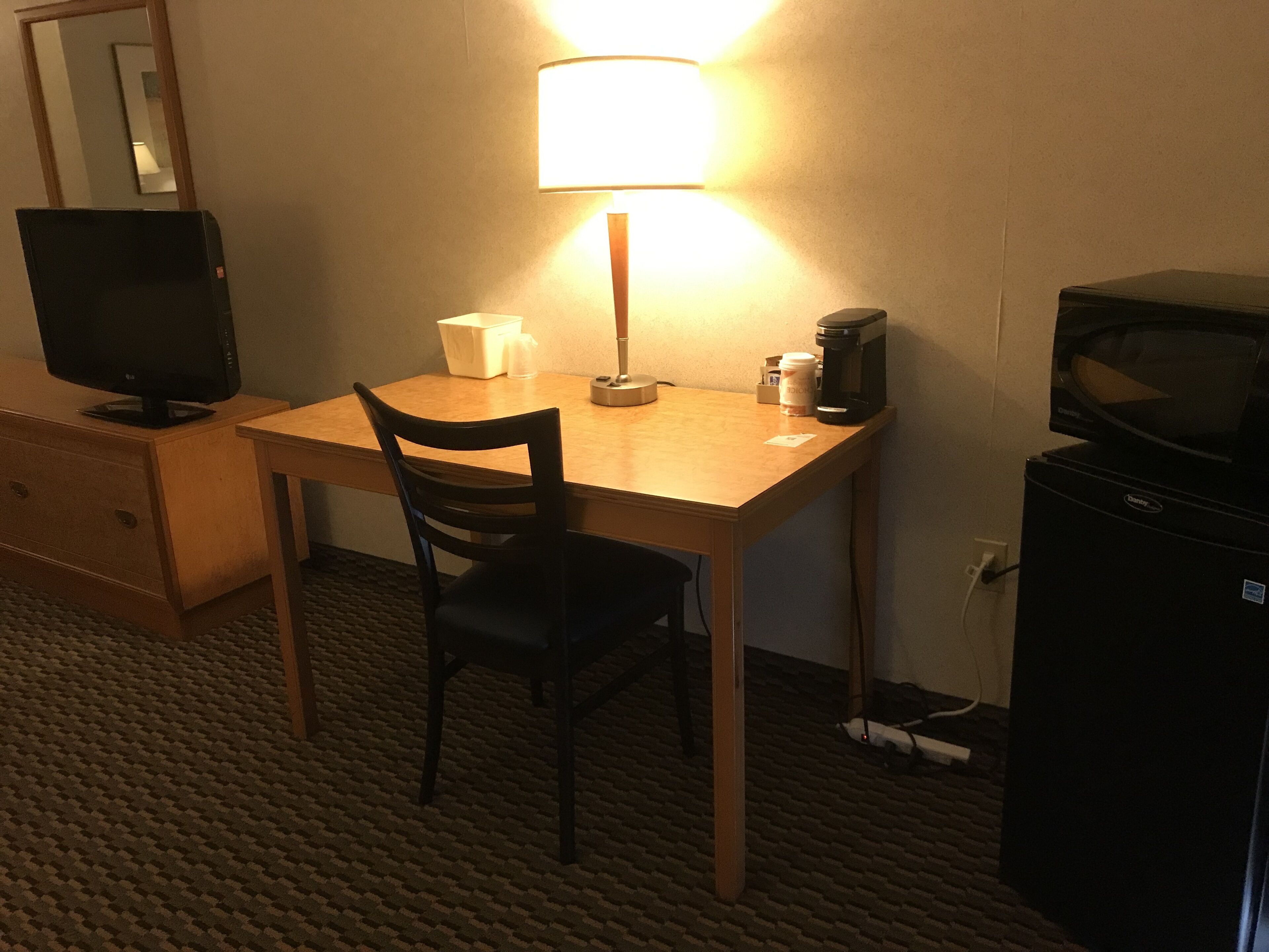 A hotel room with a wooden desk, a chair, a television, a lamp, a coffee maker, and a refrigerator.