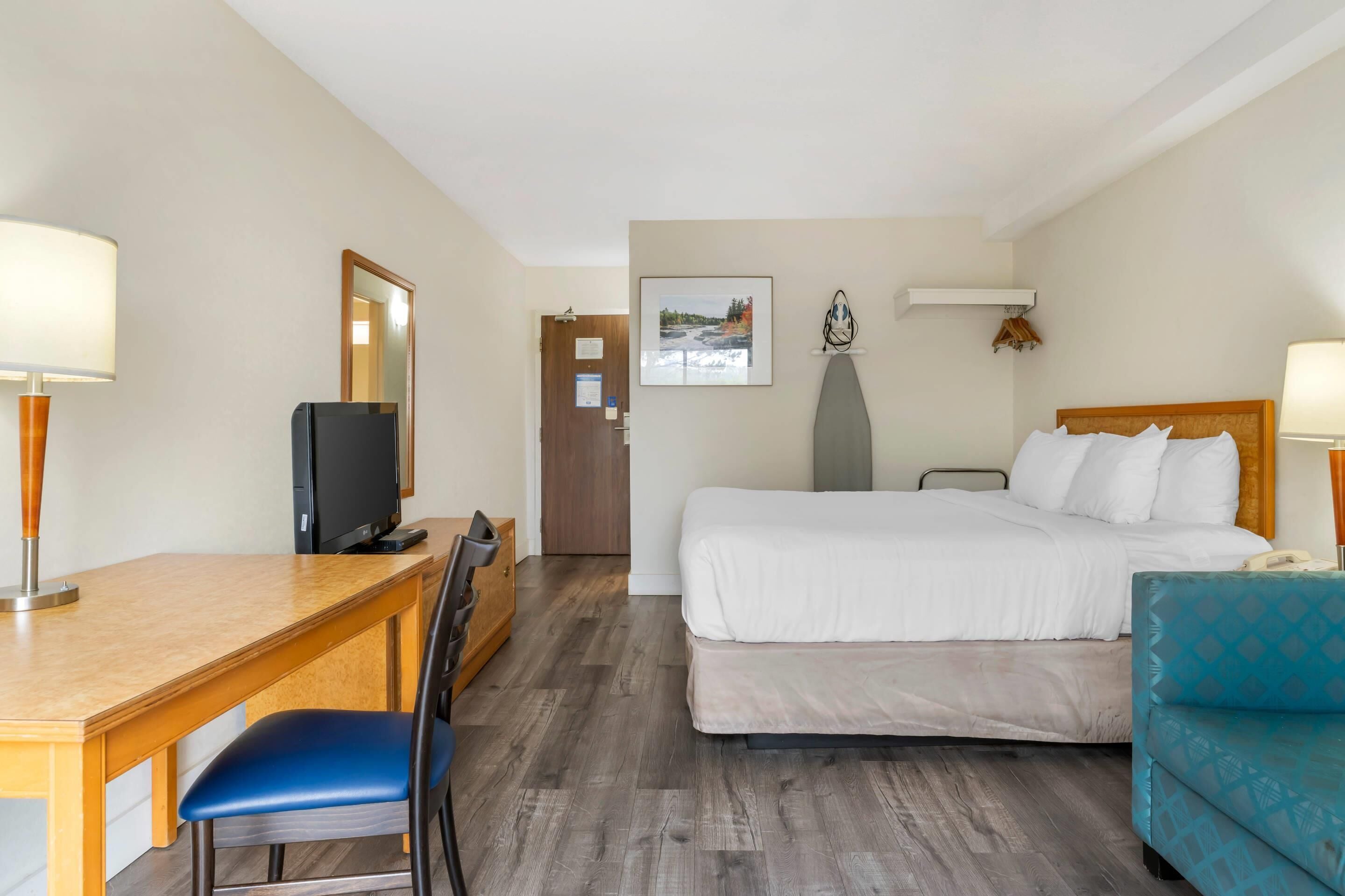 Ein Hotelzimmer mit Holzschreibtisch, Fernseher, Bett mit weißer Bettwäsche, blauem Sessel und blauem Sofa.