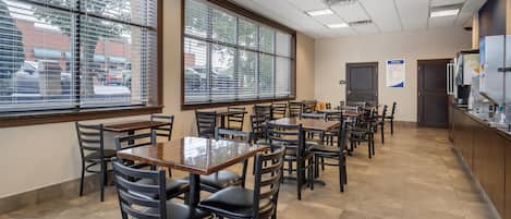 A restaurant with multiple tables and chairs, large windows with blinds, and a tiled floor.