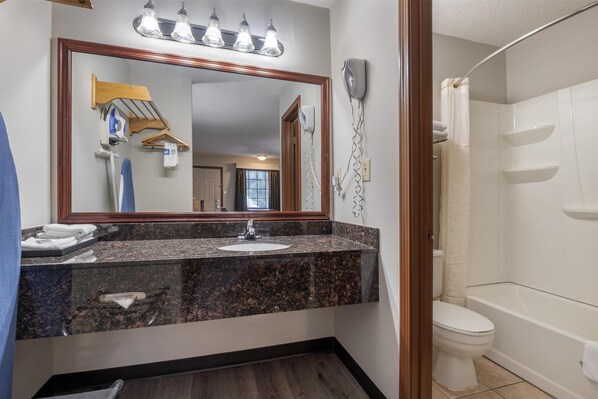 A bathroom with a granite countertop sink, a mirror with lights, a hairdryer, a toilet, and a shower area.