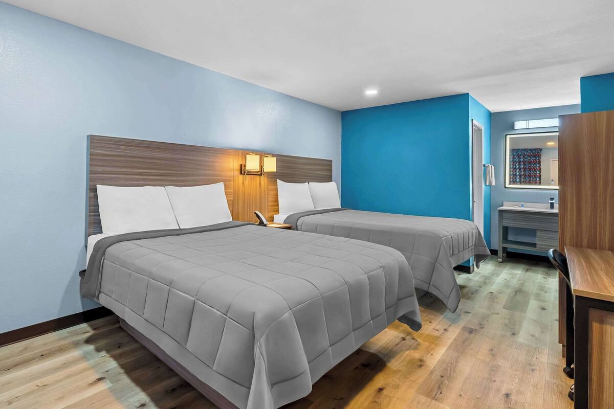 Quarto de hotel com duas camas, cabeceira de madeira, mesa de cabeceira, espelho e piso de madeira.