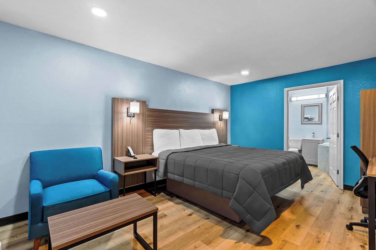 Quarto de hotel com uma cama, poltrona azul, mesa de cabeceira de madeira e uma escrivaninha com cadeira.