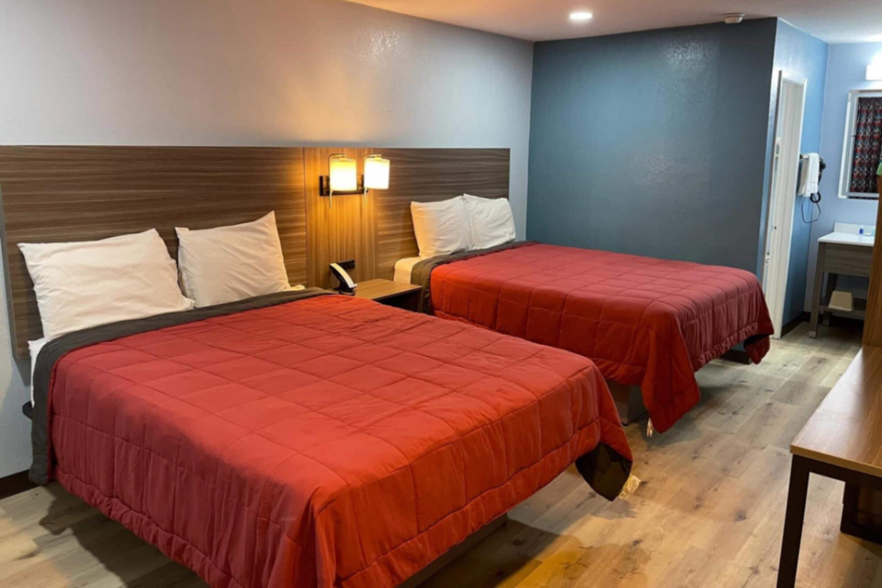 A hotel room with two beds, each with a red bedspread, a wooden headboard, and a wall-mounted lamp.