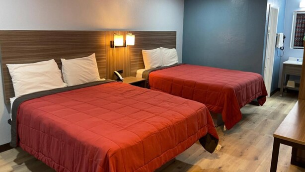 A hotel room with two beds, each with a red bedspread, a wooden headboard, and a wall-mounted lamp.