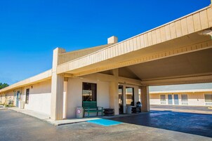 Un motel con área de estacionamiento techada, un banco verde y un bote de basura.
