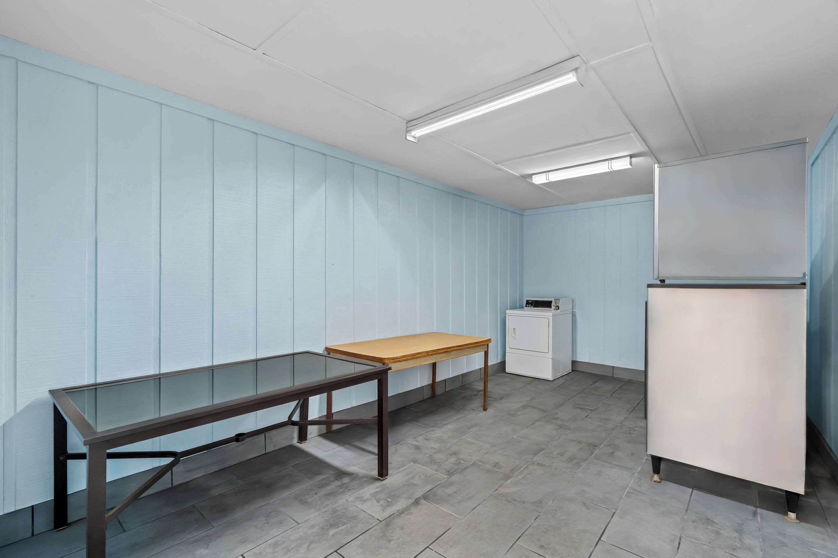 Une pièce comprenant une table à plateau en verre, une table en bois, un meuble blanc et un réfrigérateur blanc.