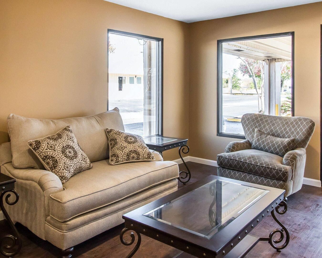 Una sala de estar con sofá y sillón, una mesa de centro de vidrio y dos ventanas con vistas al exterior.