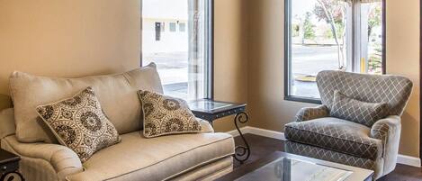 Una sala de estar con sofá y sillón, una mesa de centro de vidrio y dos ventanas con vistas al exterior.