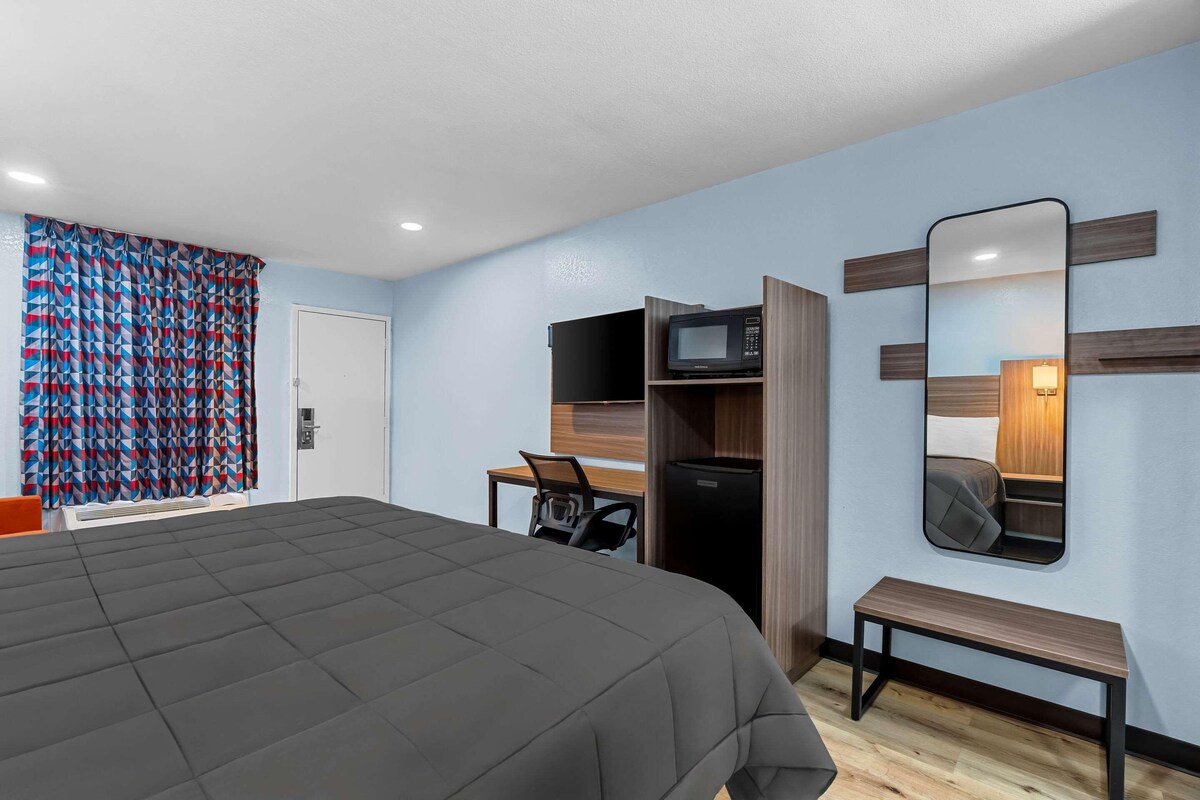 A hotel room with a bed, a desk with a chair, a microwave, a refrigerator, a bench, a patterned curtain, and a mirror.