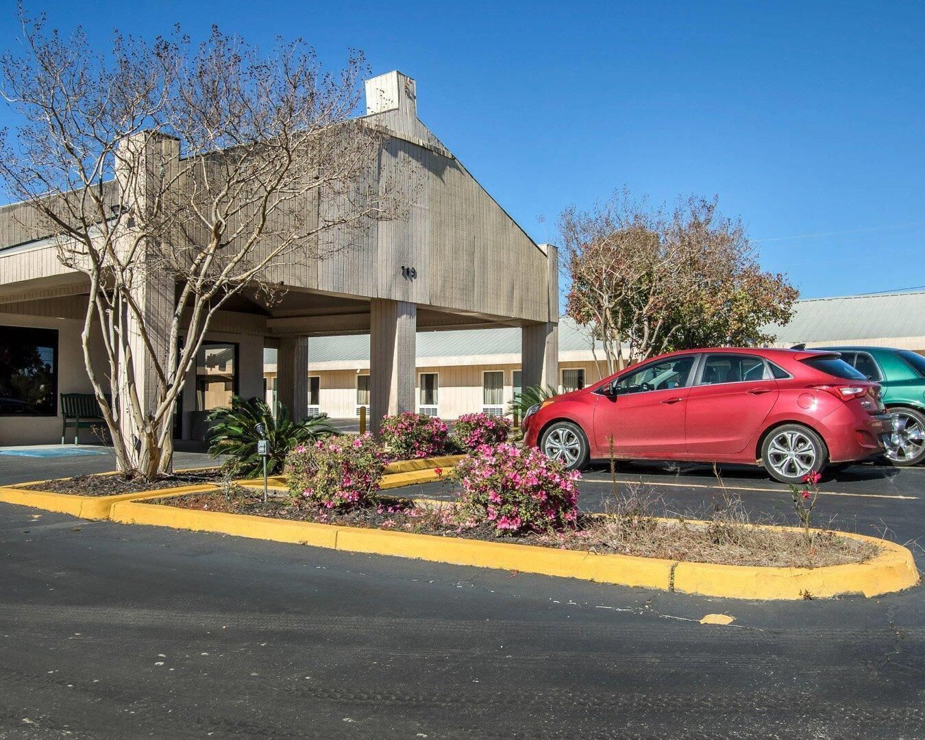 Un estacionamiento con un auto rojo estacionado frente a un edificio con el letrero '119'. Se observan bordillos amarillos y algunas plantas con flores.