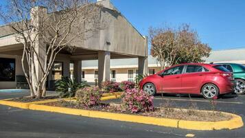 Un estacionamiento con un auto rojo estacionado frente a un edificio con el letrero '119'. Se observan bordillos amarillos y algunas plantas con flores.