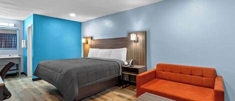 A hotel room with a bed, a desk, a chair, and a nightstand with a lamp.