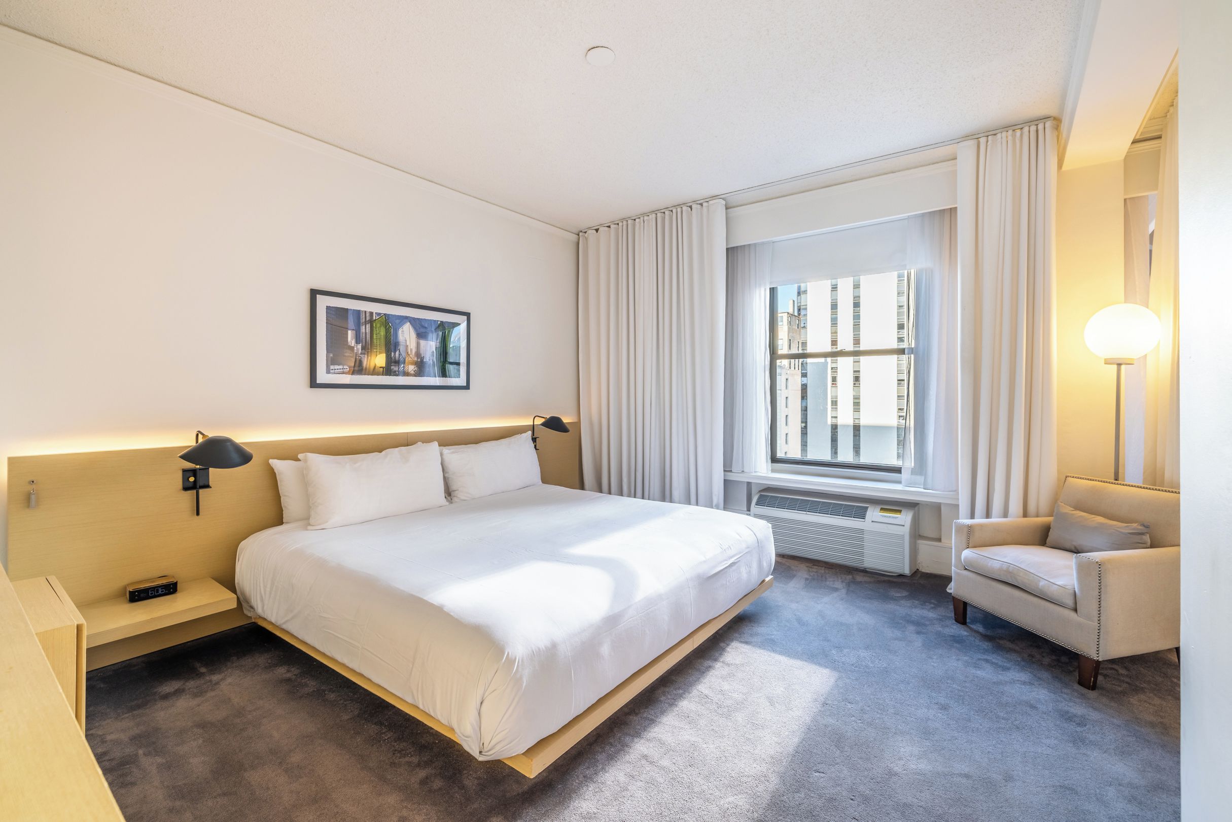Una habitación de hotel con una cama grande, una silla, una ventana con cortinas, una lámpara de pared y un cuadro enmarcado.