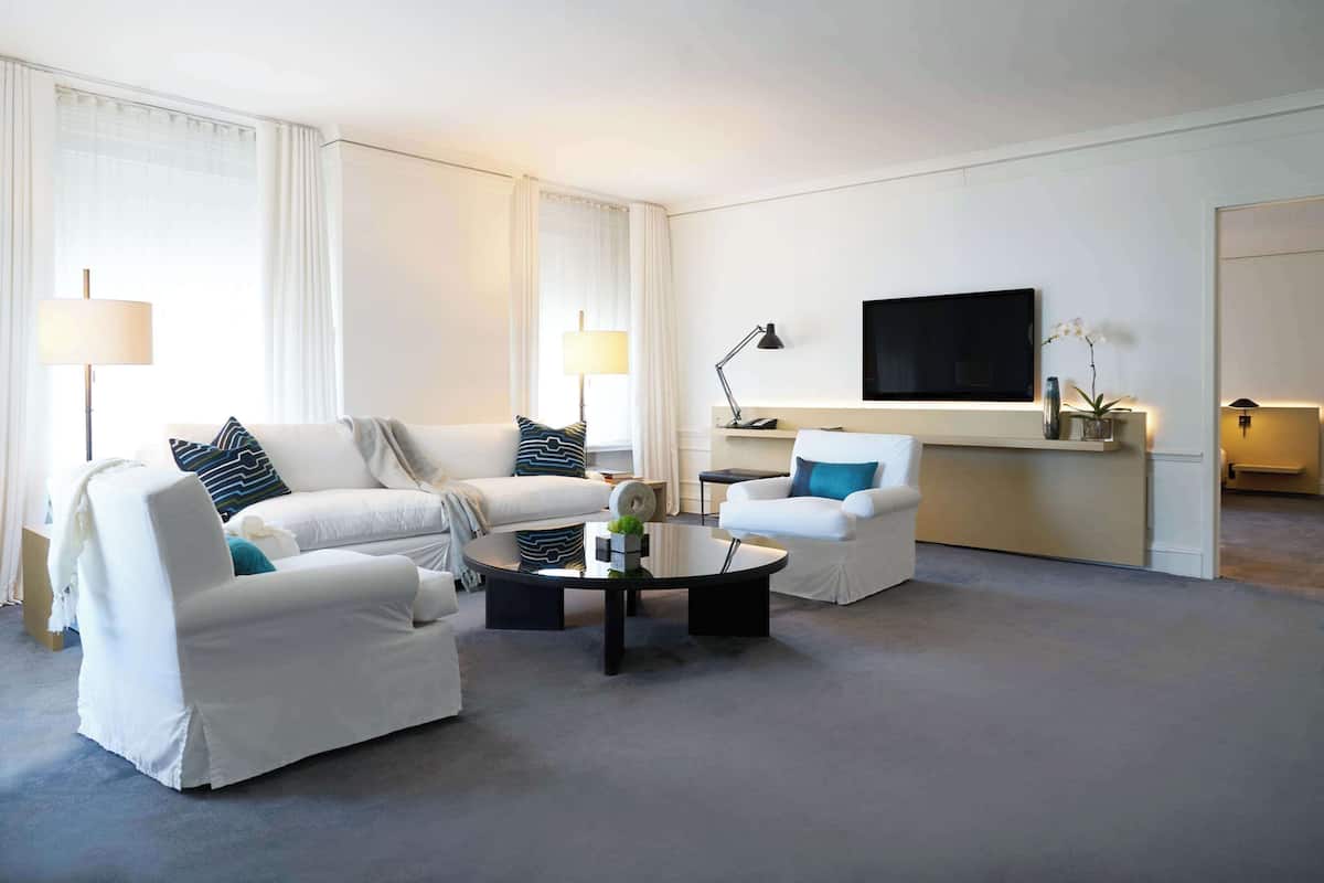 A modern hotel room with a white sofa set, a round coffee table, a flat-screen TV mounted on the wall, and a small side table with a lamp.