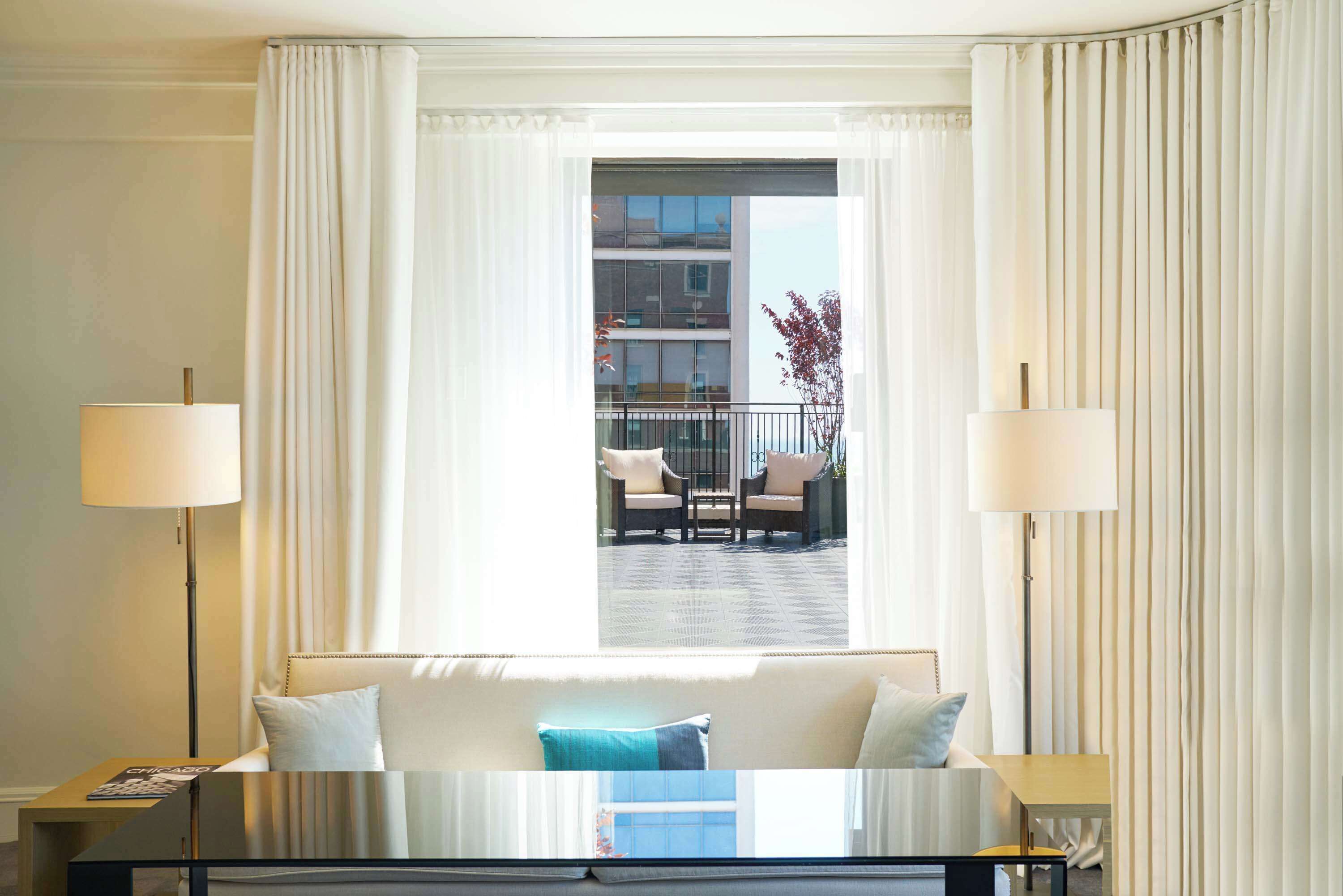 A hotel room with a large window, a bed with a blue pillow, two bedside lamps, and a view of a balcony with outdoor furniture.